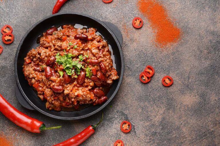 A black bowl of meat chili seen from above on a stone surface and surrounded by fresh hot peppers.