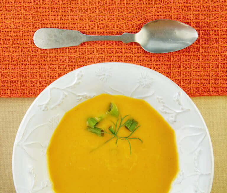 A bowl of creamy squash soup garnished with fresh herbs and green onions, served on a decorative white plate with a vintage spoon on an orange placemat.