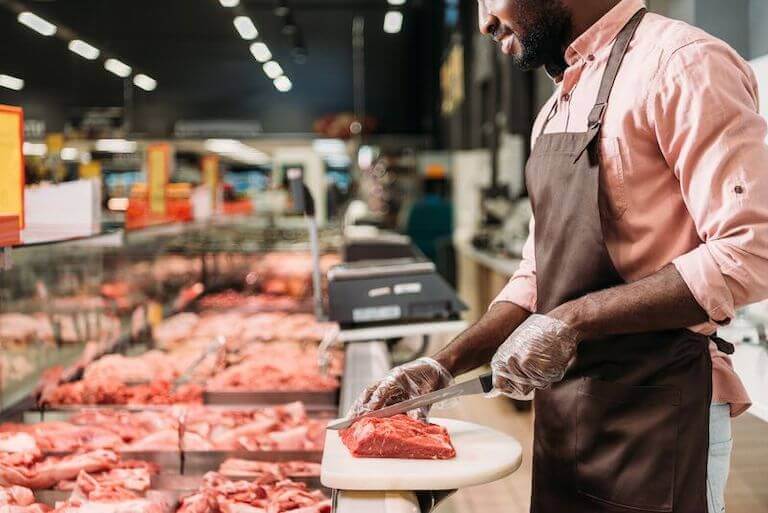 A butcher trimming meat in front of a display of a variety of meat in a retail grocery.