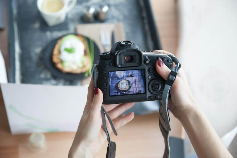 A camera in someone’s hands frames up an image of a plate of food sitting on a table near a bright window.
