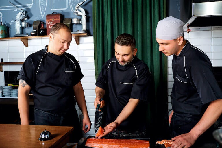 A Chef holds a piece of fish and a knife in his hand as two other chefs watch him.