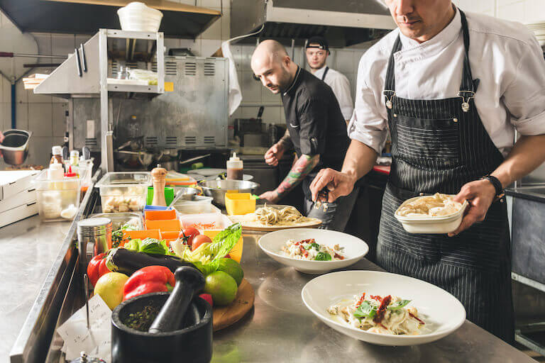 Several chefs in aprons work side-by-side in a commercial kitchen plating colorful, vegetable-forward dishes.