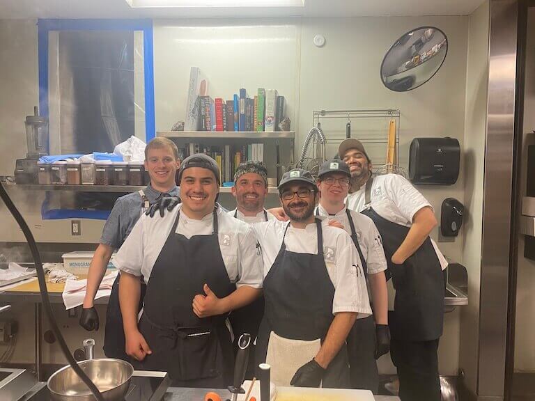 A group of six prep chefs, including Michael Fields, in chef’s whites stand smiling in a commercial kitchen.