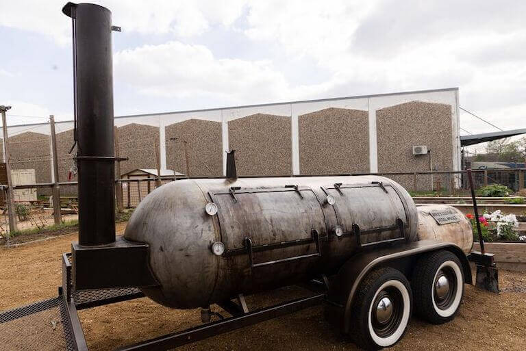 A large, metal, cylindrical barbecue smoker sits outdoors on wheels at the Escoffier Austin campus.