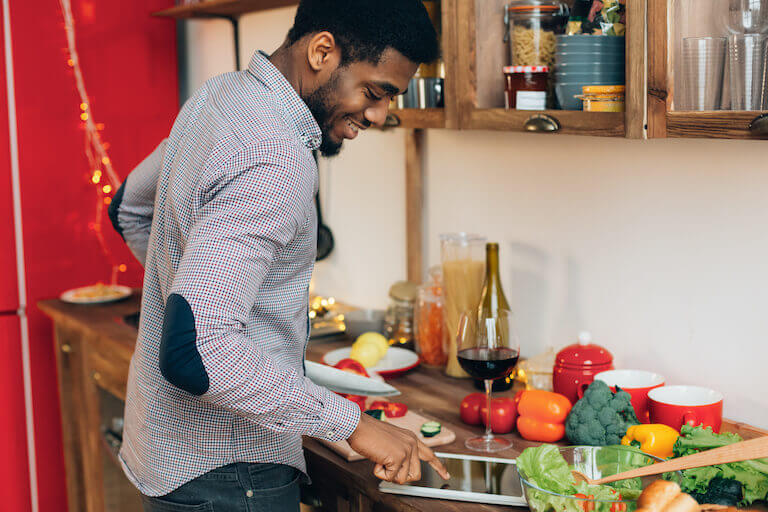 A person is reading a recipe on a tablet while standing at a counter cutting vegetables for a meal.