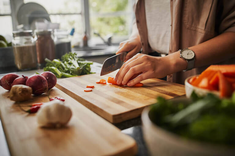 A pair of hands chops celery on a wooden cutting board. Tomatoes and herbs are already chopped and on the board.