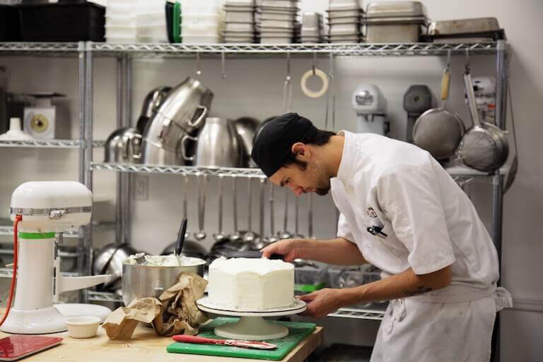 A baker in a commercial kitchen leans over a cake that they have just started frosting and decorating.
