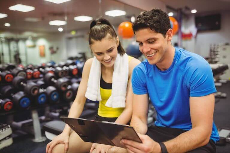 A person and a trainer meeting at a gym and looking at a tablet with weights in the background.