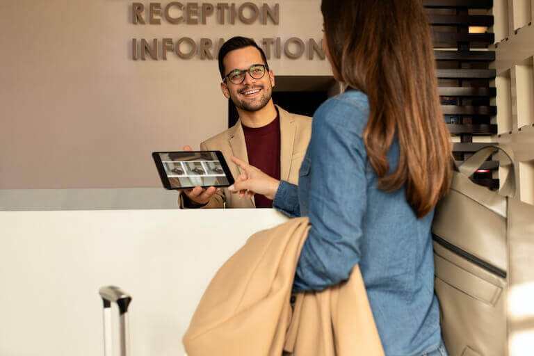 A person behind the front desk of a hotel smiles and shows a guest, whose back is to the camera, a tablet with room choices.