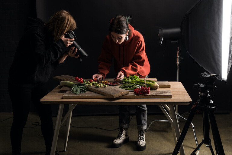 A photographer in black clothes frames up a platter of fresh vegetables being arranged by a food stylist in a red hoodie.