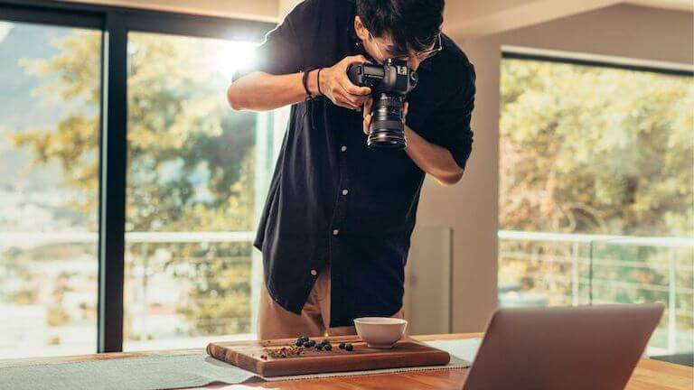 A photographer stands above a wooden cutting board with a white bowl and berries on it, setting up an overhead shot.
