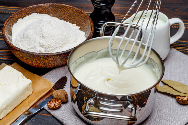 A white béchamel sauce in a pan with whisk, flour, and ingredients surrounding it.