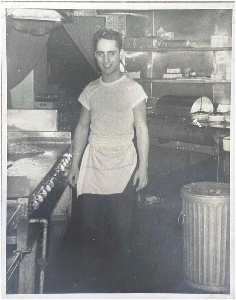 A young Joey Vento stands smiling in the Geno’s Steaks kitchen wearing a white t-shirt and a white apron in a black and white photograph.
