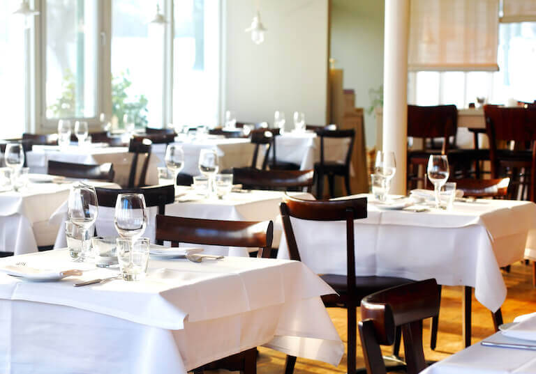 An empty restaurant featuring tables with white tablecloths and full place settings.