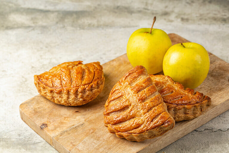 Golden apple turnovers resting on a wooden board next to fresh apples, highlighting their flaky crust and sweet filling.