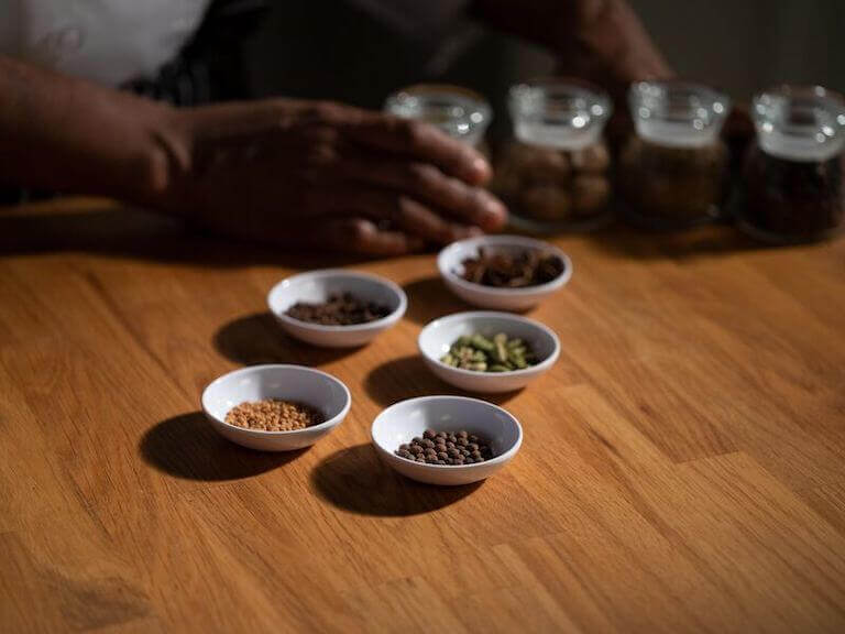 Assorted spices in small bowls arranged on a wooden table, featuring whole spices like cumin seeds, cardamom pods, and cloves, highlighting ingredients used in flavor-enhancing techniques.