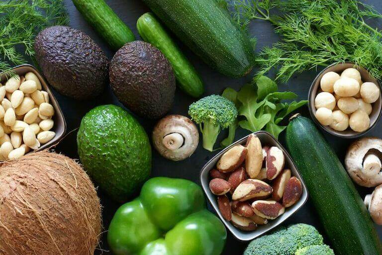 An assortment of healthy vegetables, like avocados, broccoli, mushrooms, and zucchini are arranged artfully on a table.