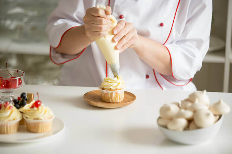 A chef pipes buttercream frosting onto a vanilla cupcake.