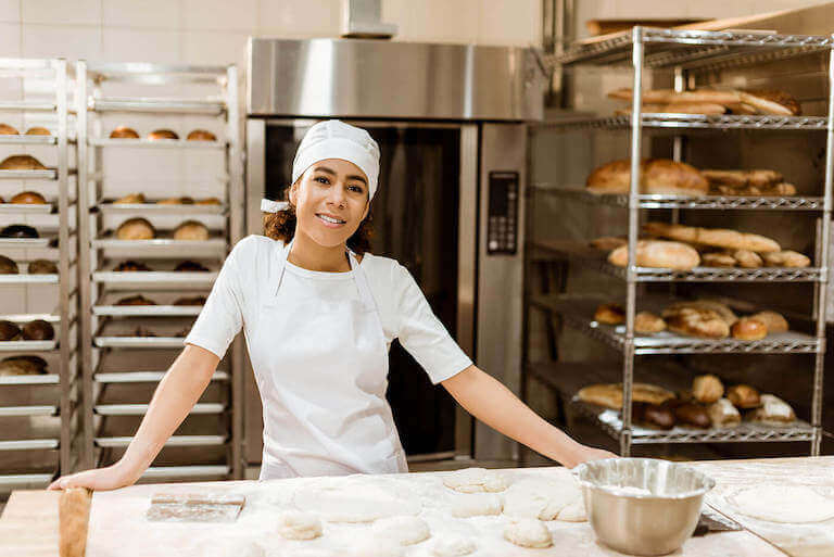 Baker standing at a table with fresh dough in front of her and racks of baked bread in the background.