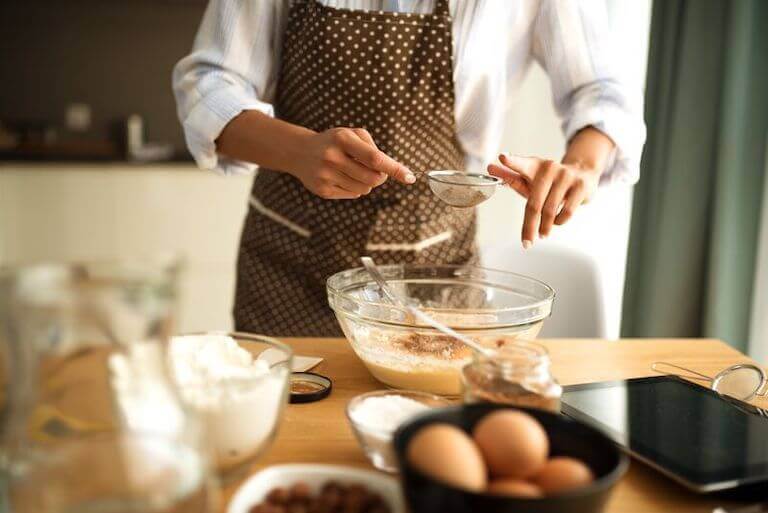 A person wearing a brown apron with white polka dots sifts ingredients into a large glass mixing bowl sitting on a table surrounded by baking ingredients.