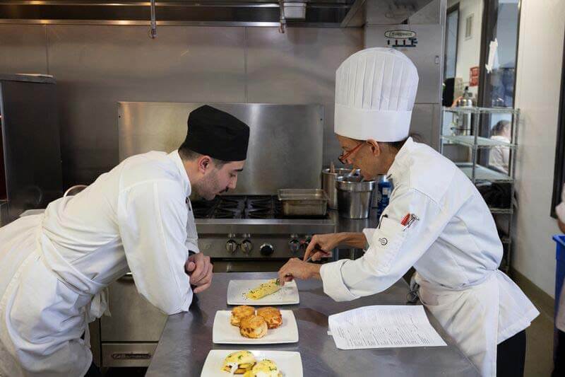 A chef observes closely as another chef plates an omelette on a white plate, next to a plate of biscuits and Eggs Benedict.