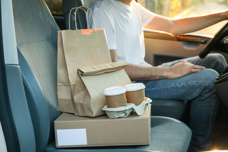 To-go boxes, bags, and cups sit in the passenger seat of a car, with a driver behind the wheel.