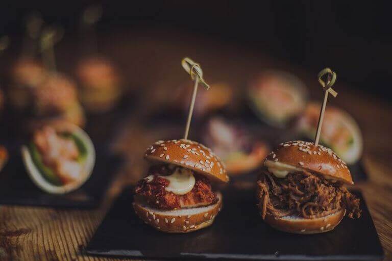 Two gourmet mini burgers with sesame seed buns, topped with sauce and skewered with picks, plated on a dark wooden surface with blurred appetizers in the background.