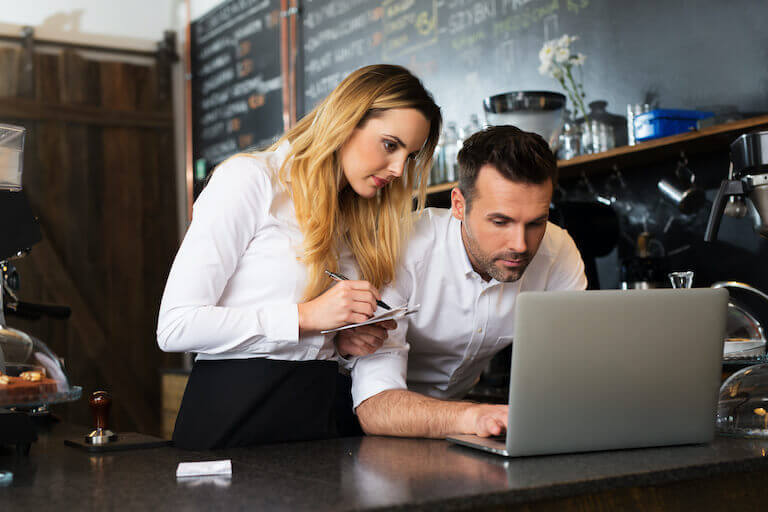 Cafe manager and waitress take notes as they look at a laptop.