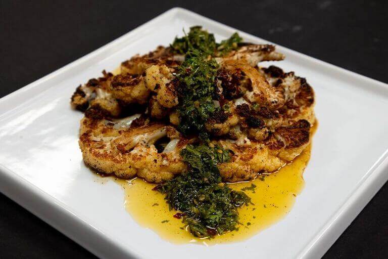 Grilled slabs of cauliflower lie on a square plate, garnished with parsley.