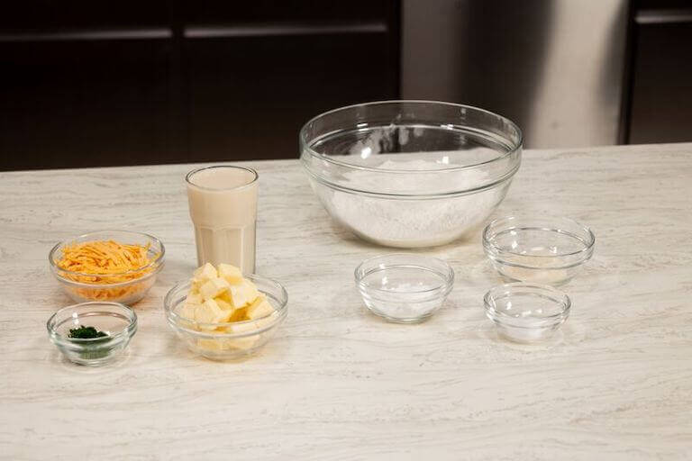 Glass bowls containing shredded cheese, flour, and other pre-measured ingredients for making biscuits arranged on a countertop.