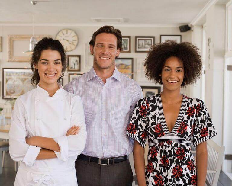 Three people stand in a small cafe, facing the camera and smiling.