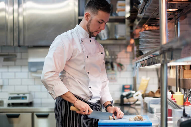 A chef stands at a counter in a commercial kitchen and chops shrimp on a large cutting board.