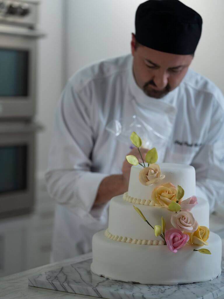 A pastry chef in a white uniform and black cap carefully decorates a three-tiered white cake adorned with delicate sugar flowers in a professional kitchen.