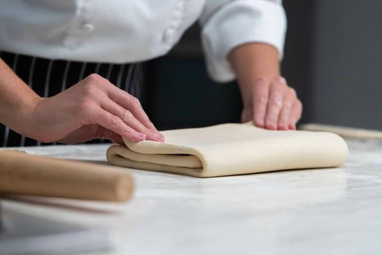 Chef folding croissant dough by hand on a floured surface in a professional kitchen, preparing laminated dough for baking.