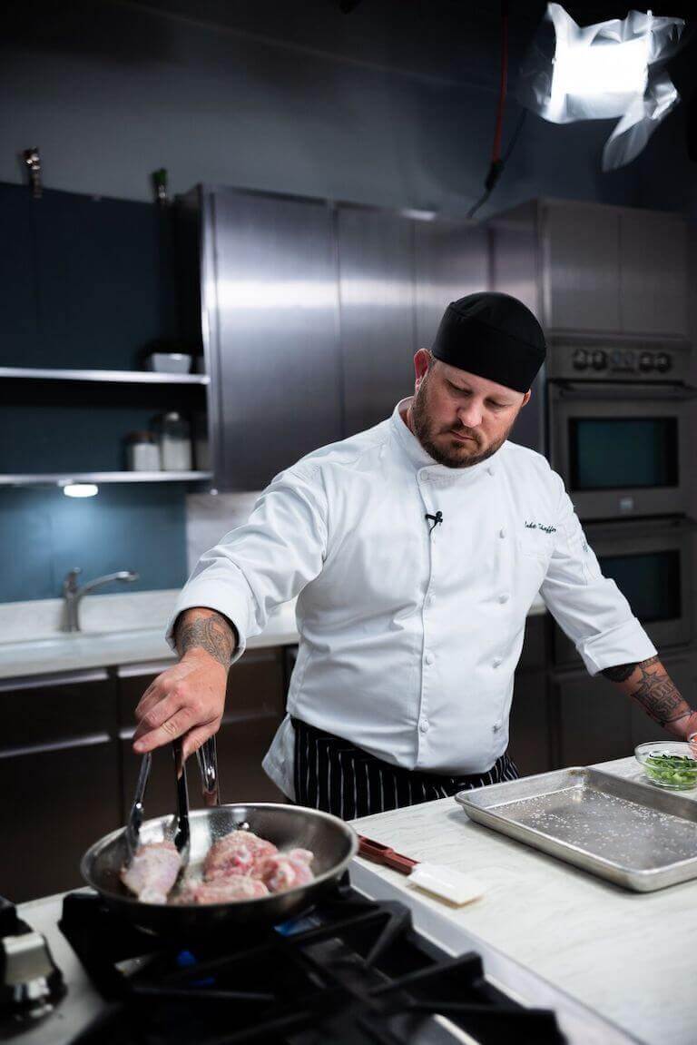 A chef in a white jacket and black cap carefully sears chicken in a pan with tongs in a modern, professional kitchen.