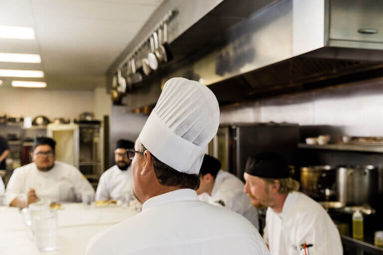A chef instructor is in the foreground with back to the camera, speaking to a group of students who are seated around a counter in a professional kitchen in the background.
