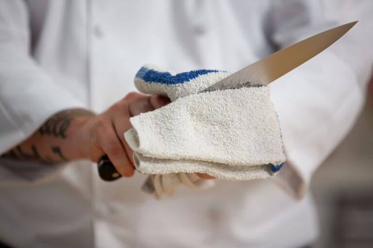 A close-up of a chef wiping a chef’s knife with a kitchen towel.