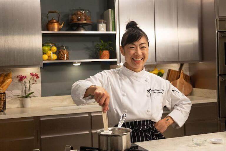 Chef Erin Kanagy-Loux smiling while stirring sauce in metal pot during online Pastry Arts instruction.