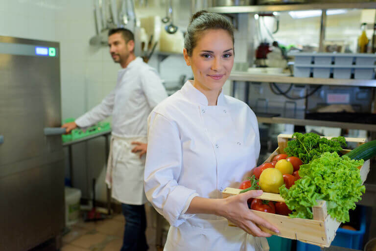 Chef standing in a kitchen holding a crate of fresh vegetables