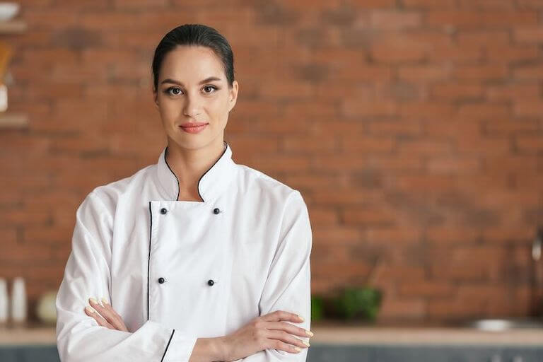 A person in a chef's coat stands in a kitchen facing the camera with their arms crossed.