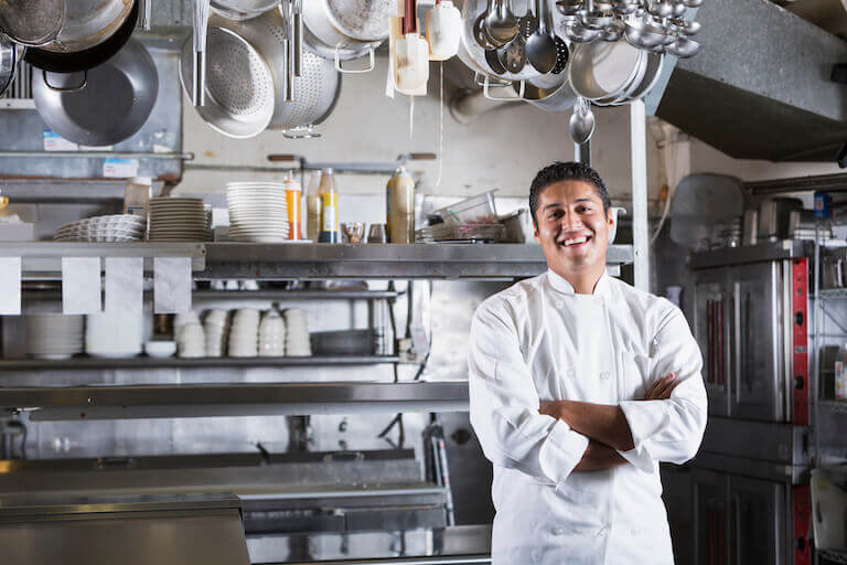 A smiling Chef de Cuisine standing with arms crossed in a commercial kitchen, surrounded by pots and pans hanging above him.