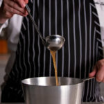 Chef using ladle to pour chicken stock broth into metal pot