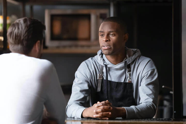 Chef in apron sitting at table having conversation with mentee in restaurant setting