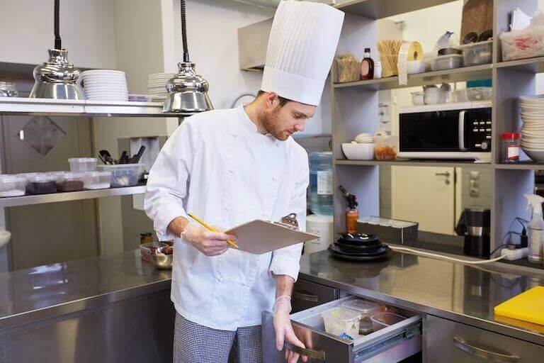 A person in a chef’s uniform holds a clipboard in one hand while opening a refrigerator drawer to check inventory in a kitchen.
