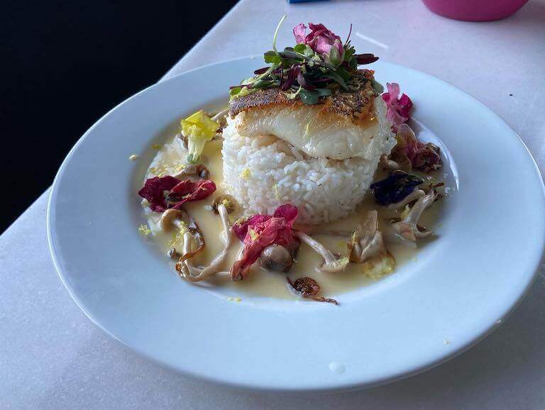 An artfully plated piece of Chilean sea bass and a mound of rice sits in a silky gravy sprinkled with flowers, mushrooms, and herbs.