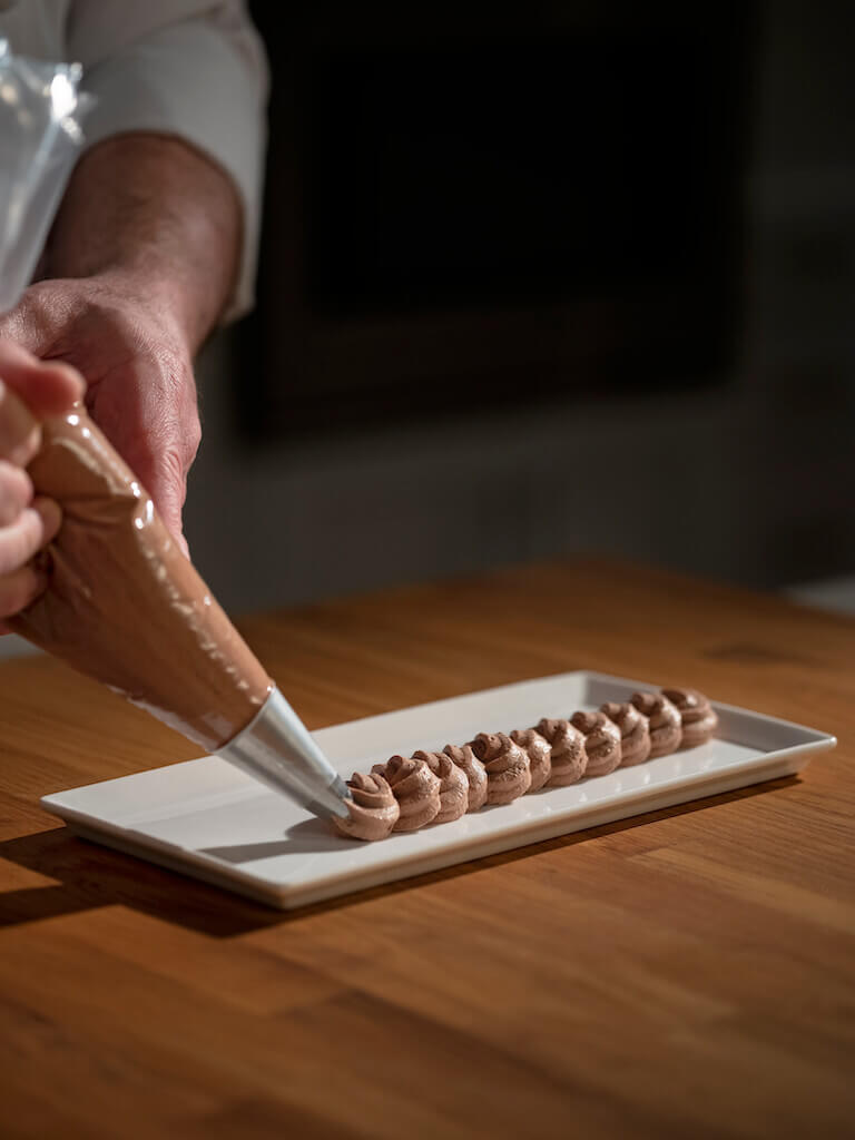 Pastry Chef using a piping bag to carefully apply chocolate mousse onto a white rectangular plate.