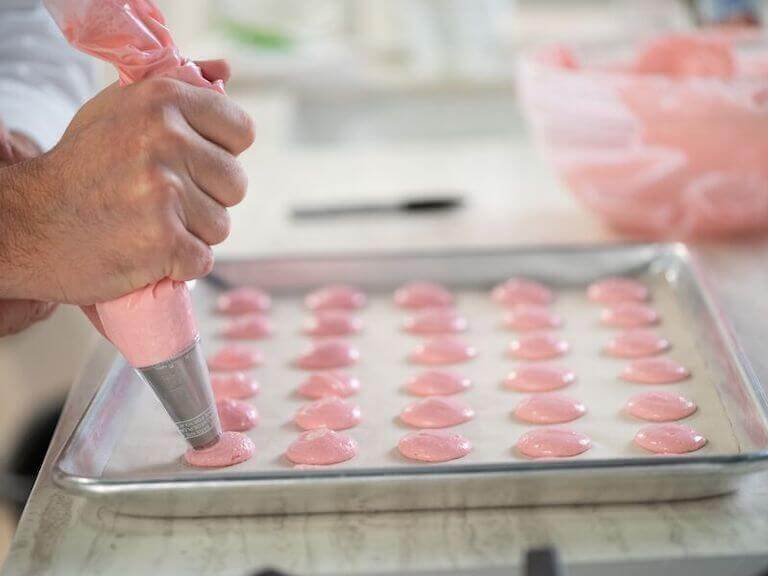 A close-up of a baker's hands piping pink macaron batter onto a lined baking sheet in a bright kitchen.