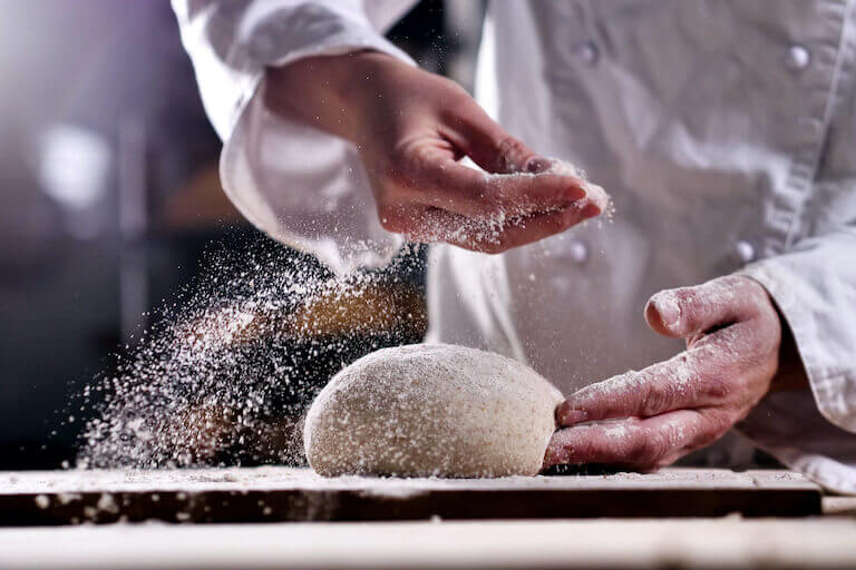 Closeup of a pair of hands sprinkling flour over a mound of dough that is sitting on a cutting board.