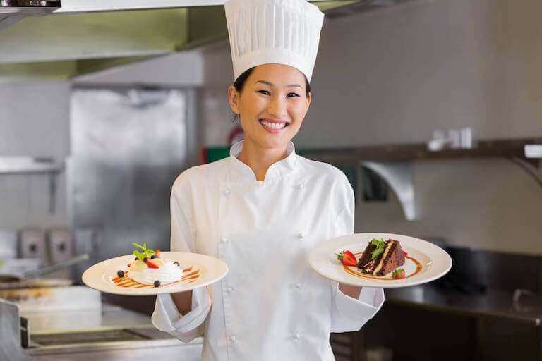 A smiling pastry chef in a white uniform and tall chef’s hat holds two plates with elegantly plated desserts in a professional kitchen.