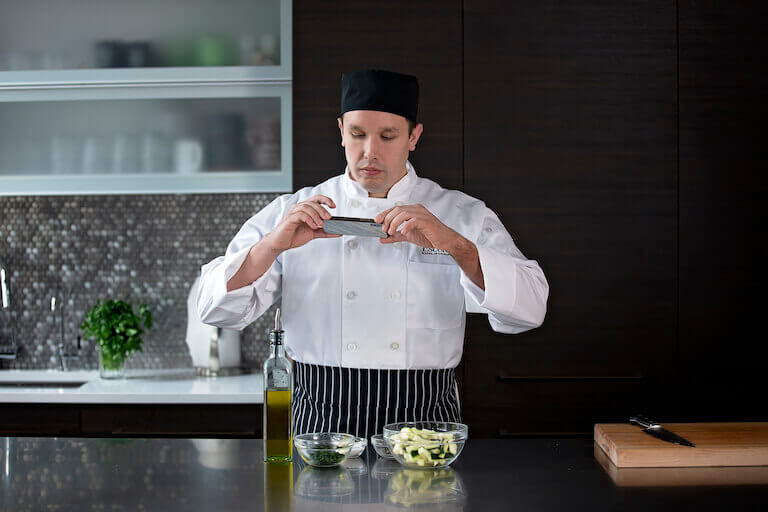 Online culinary student in chef’s whites and apron taking a photo of their mise in place in home kitchen, representing the flexibility of online culinary education.